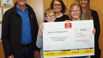 Im Vordergrund halten zwei Kinder einen Scheck über 48.000 € hoch, im Hintergrund stehen drei Erwachsene.