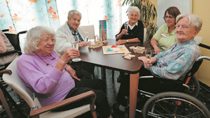 Fünf ältere Frauen, die gemeinsam an einem Tisch sitzen, ein Spiel spielen und Sekt trinken.